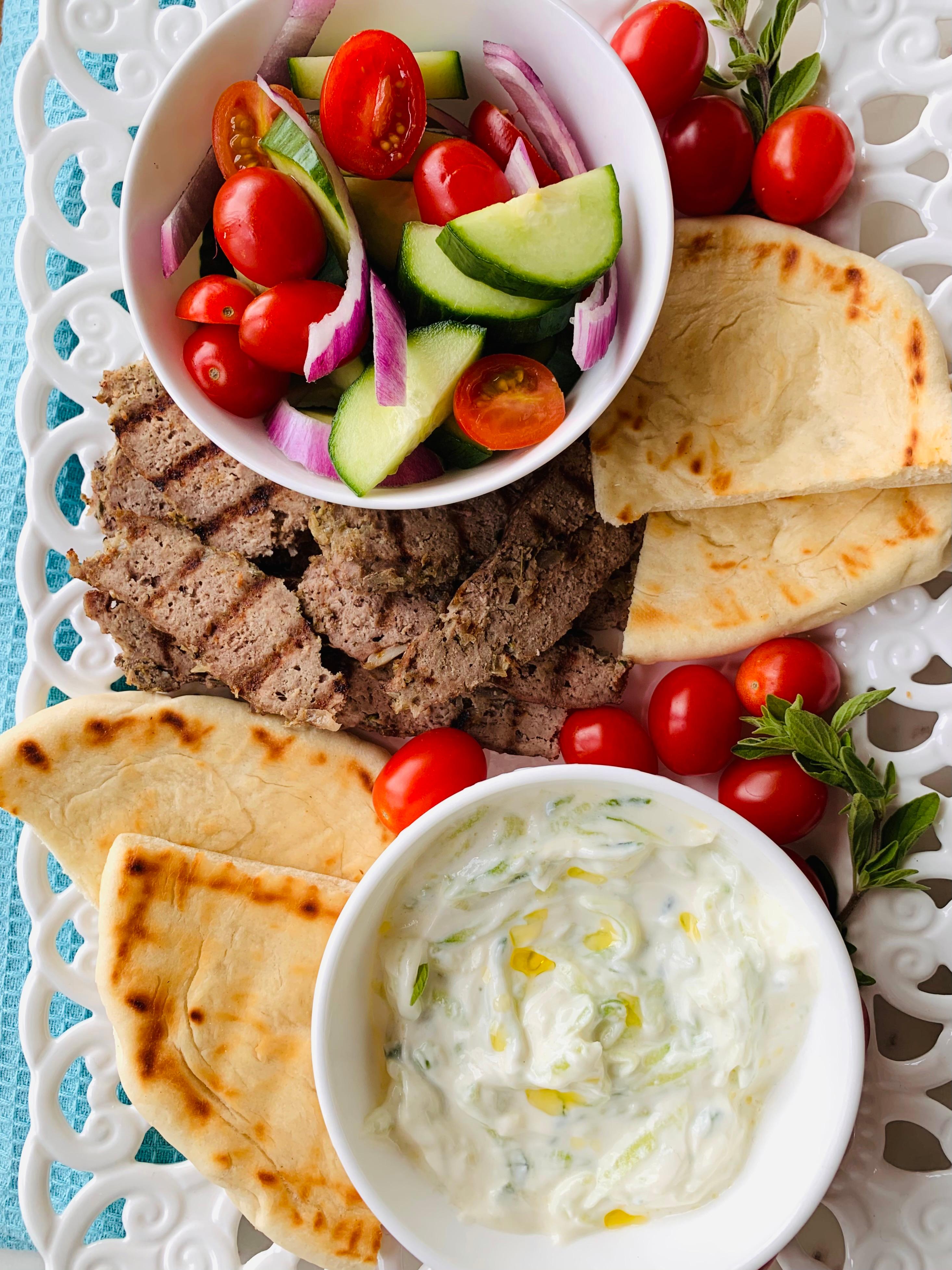 assorted greek ingredients on a wooden table, including lamb meat, pita bread, cucumbers, and yogurt