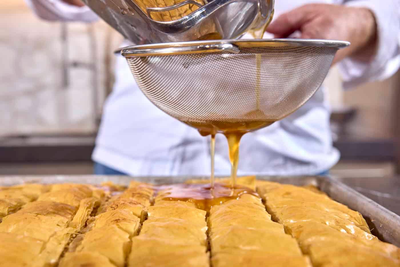 close-up shot of a hand pouring syrup over a tray of freshly baked baklava