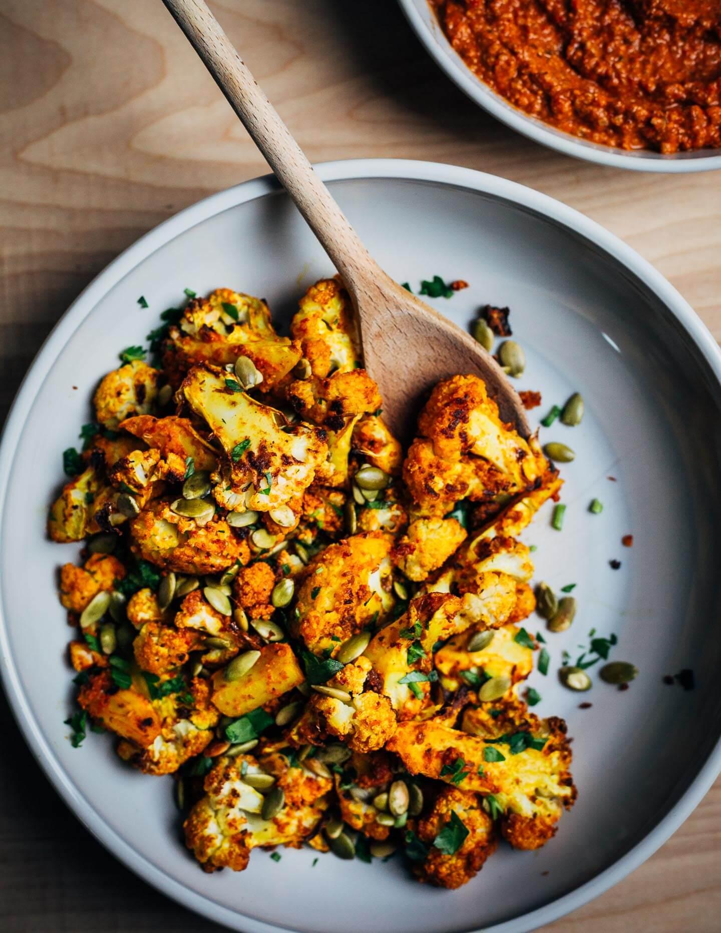 A rustic wooden table with a roasting pan of perfectly roasted potatoes, cauliflower florets, and a vibrant bowl of romesco sauce.