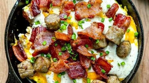 A hearty breakfast bowl with golden tater tots, scrambled eggs, cheese, and bacon, garnished with chives, on a rustic wooden table.