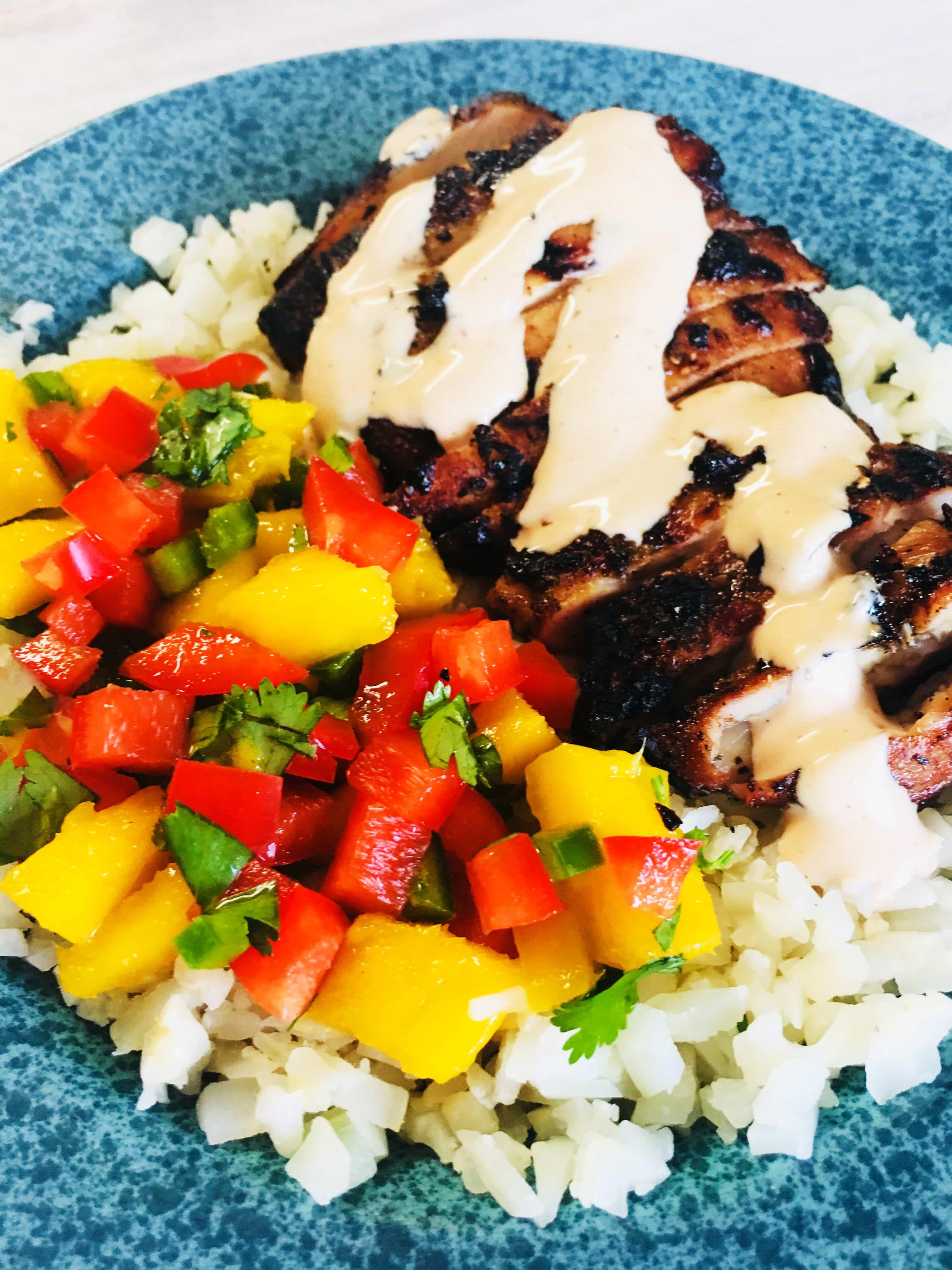 A vibrant bowl of cauliflower rice topped with pieces of Caribbean jerk chicken, garnished with fresh cilantro and a lime wedge