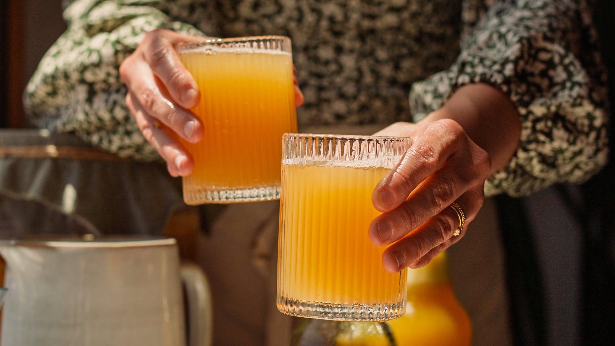 a person holding a glass of Tangerine Thyme Tonic Kombucha in a sunlit kitchen