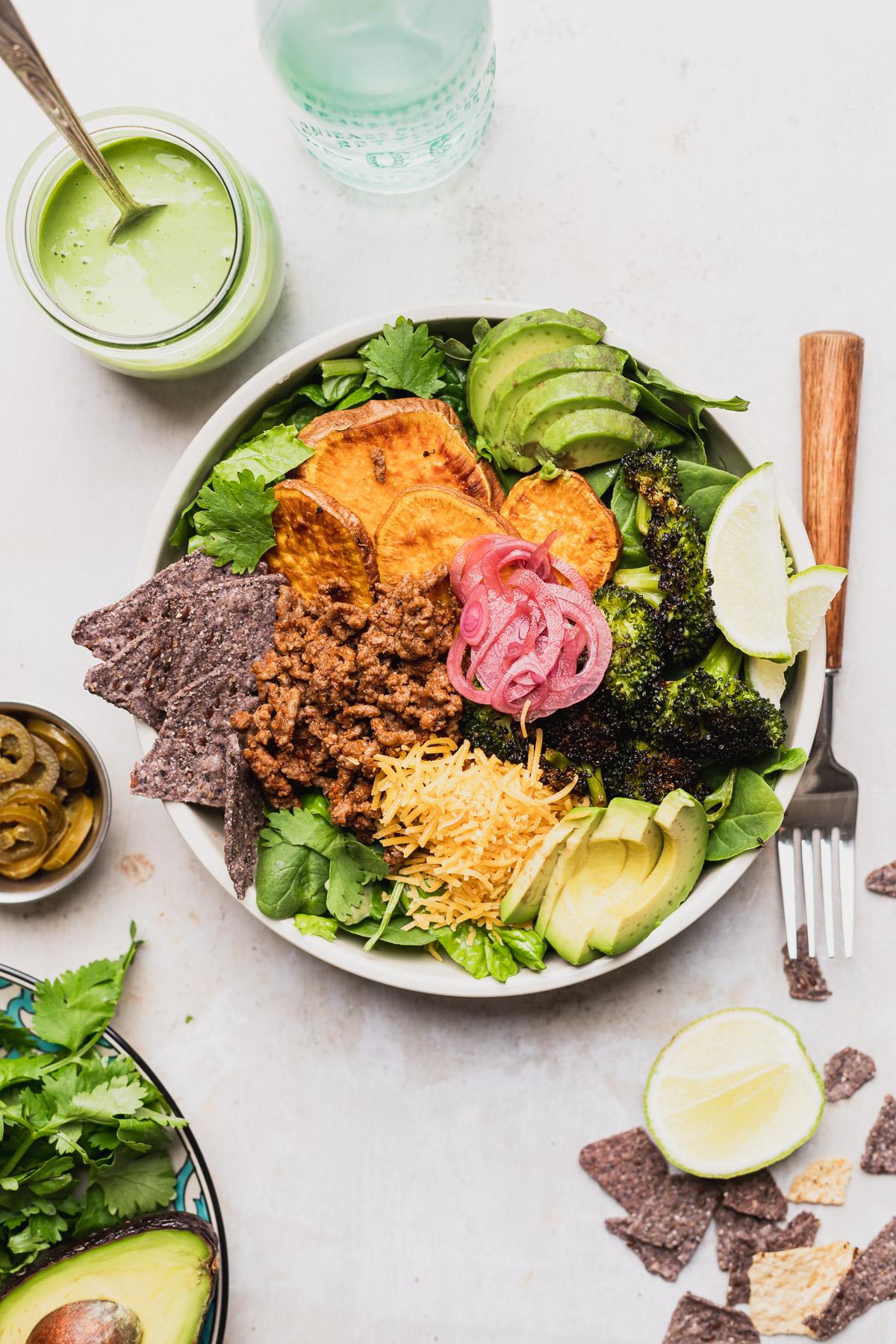 A vibrant and colorful taco salad with avocado crema, garnished with fresh cilantro and lime wedges