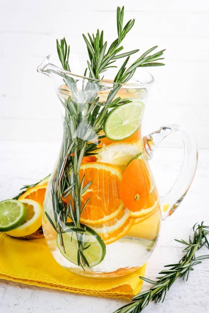 Close-up of a glass pitcher filled with rosemary citrus infused water, with lemon slices and rosemary sprigs visible