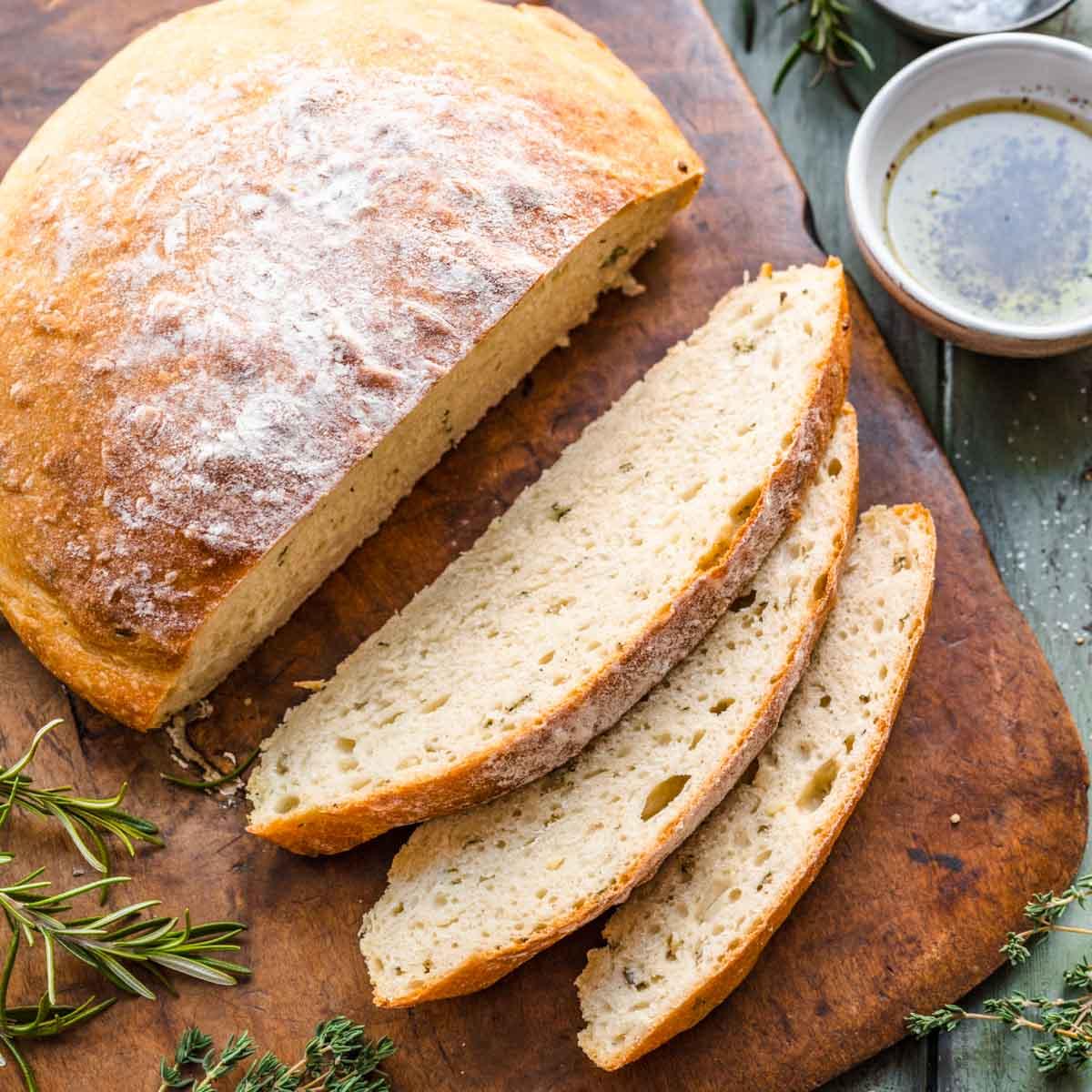 A rustic shot of a 9x13 inch baking dish of Garlic Herb Bagel Bread Pudding sitting on a wooden table, surrounded by fresh herbs and a cup of coffee.