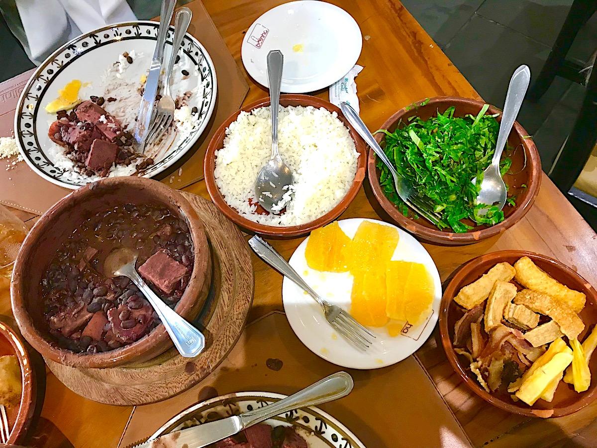 A wide shot of a table set for dinner with a large pot of Brazilian Feijoada in the center, surrounded by various side dishes.