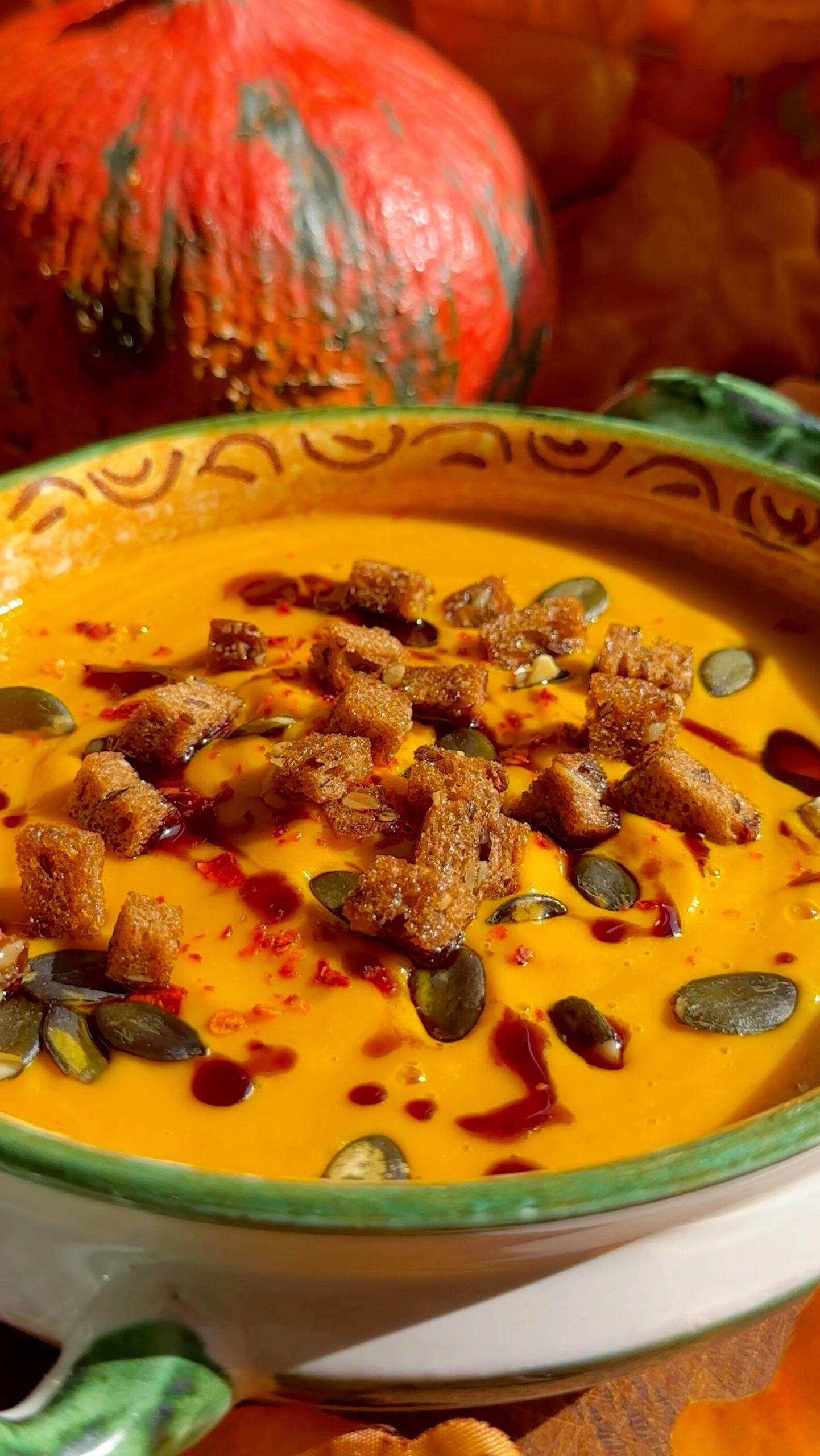 A rustic bowl of spiced pumpkin soup garnished with toasted pepitas, fall leaves in the background
