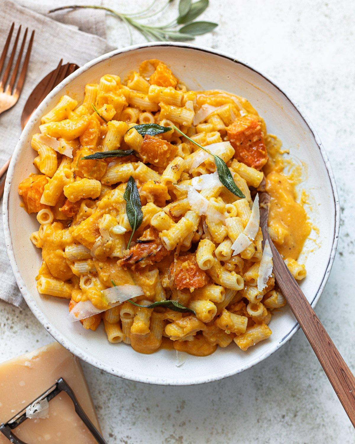 Ingredients for spiced butternut squash pasta, including butternut squash, spices, pasta, parmesan cheese, and sage on a wooden surface