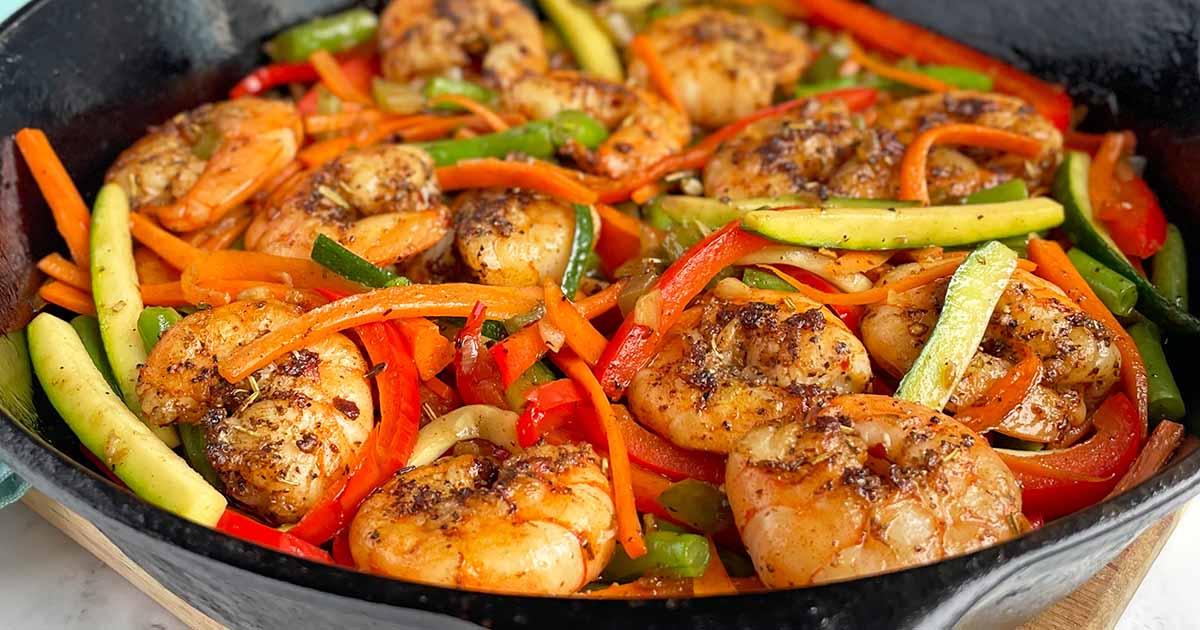 a pan with sauteed onions, bell peppers, and celery, with shrimp being added to the mix