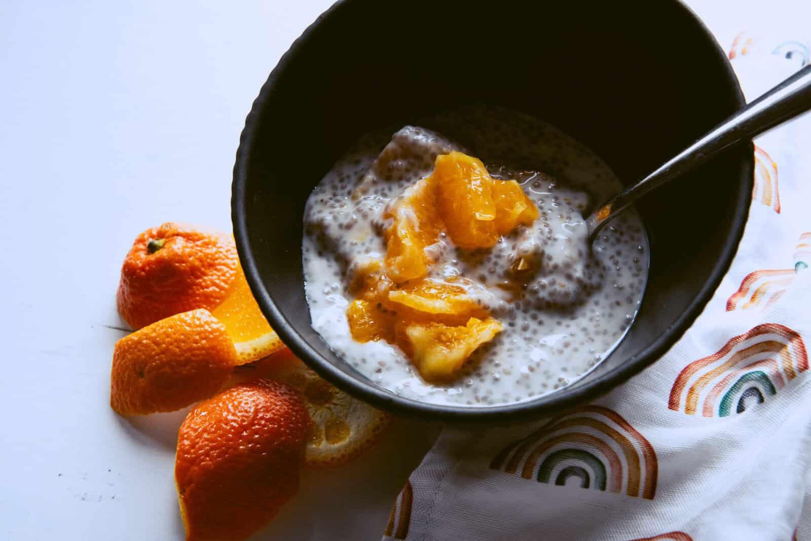 A bowl of orange blossom chia seed pudding with candied orange peel next to a bowl of fresh oranges