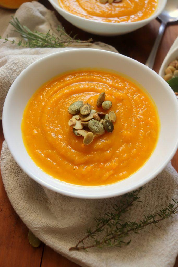 Close-up of a bowl of butternut squash soup being garnished with toasted pumpkin seeds and fresh herbs