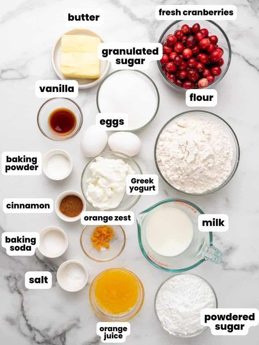 Ingredients of the cranberry orange muffins, flour, sugar, cranberries, orange zest, butter on a white marble background