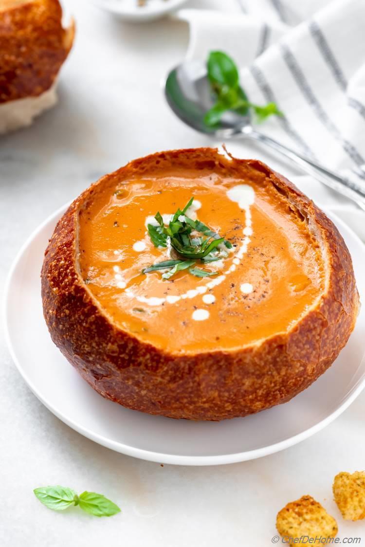 A hearty bowl of creamy tomato soup stew with a garnish of fresh basil, served with a crusty bread roll on the side, shot with warm, inviting lighting.