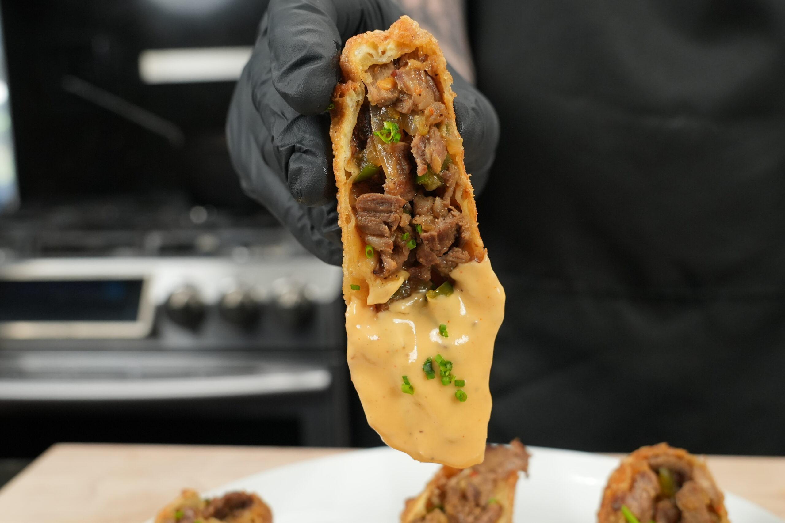 A person taking a bite of a Philly Cheesesteak Egg Roll, showing the melted cheese and savory filling.