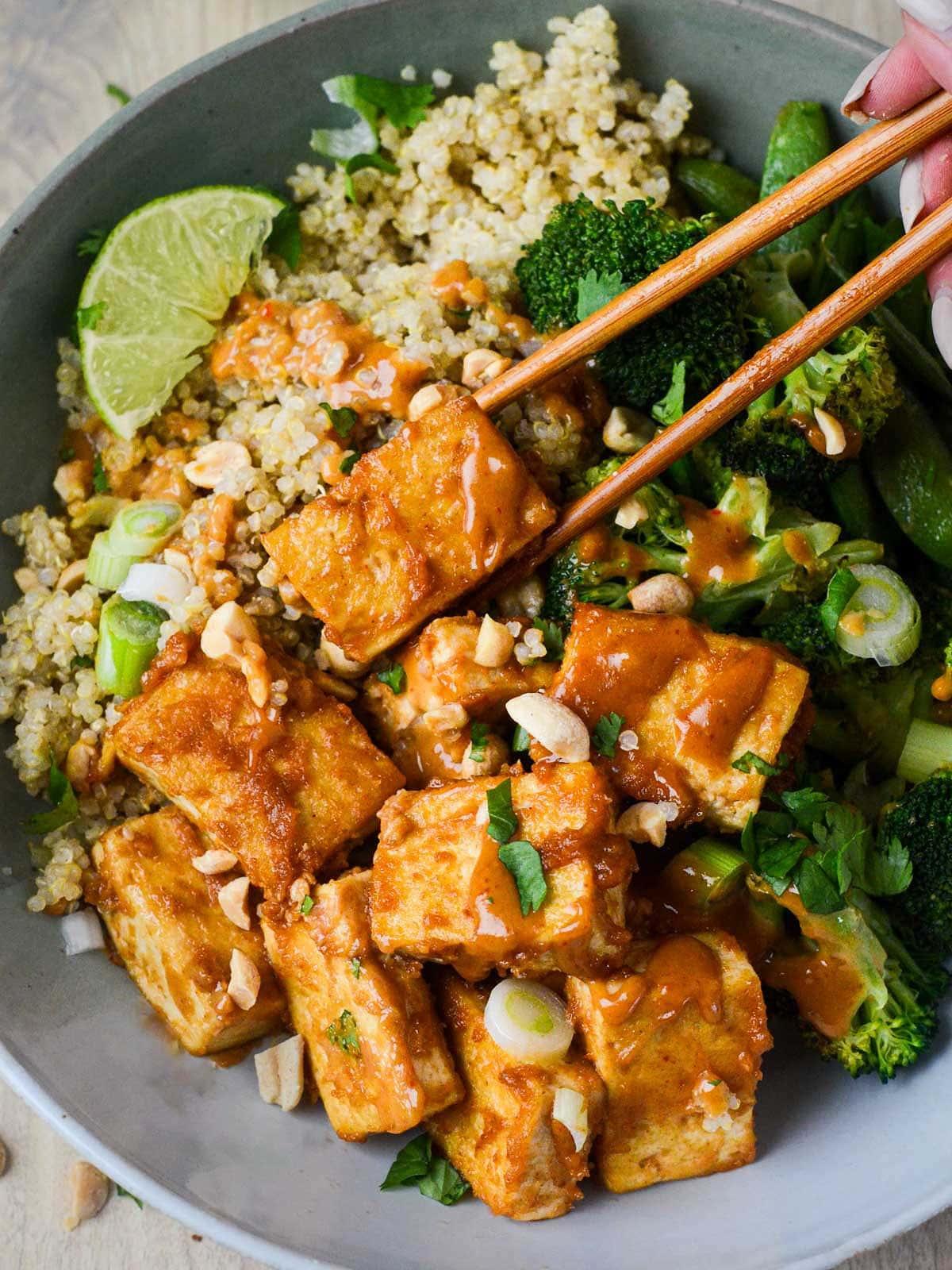 A person enjoying a bowl of quinoa salad with tofu and peanut sauce outdoors.