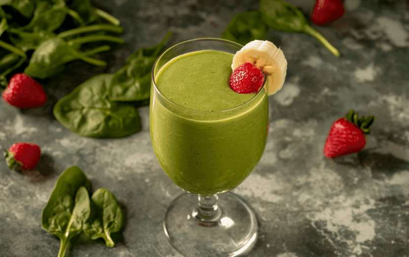 A vibrant green smoothie in a glass with ingredients like spinach, banana and berries around it.