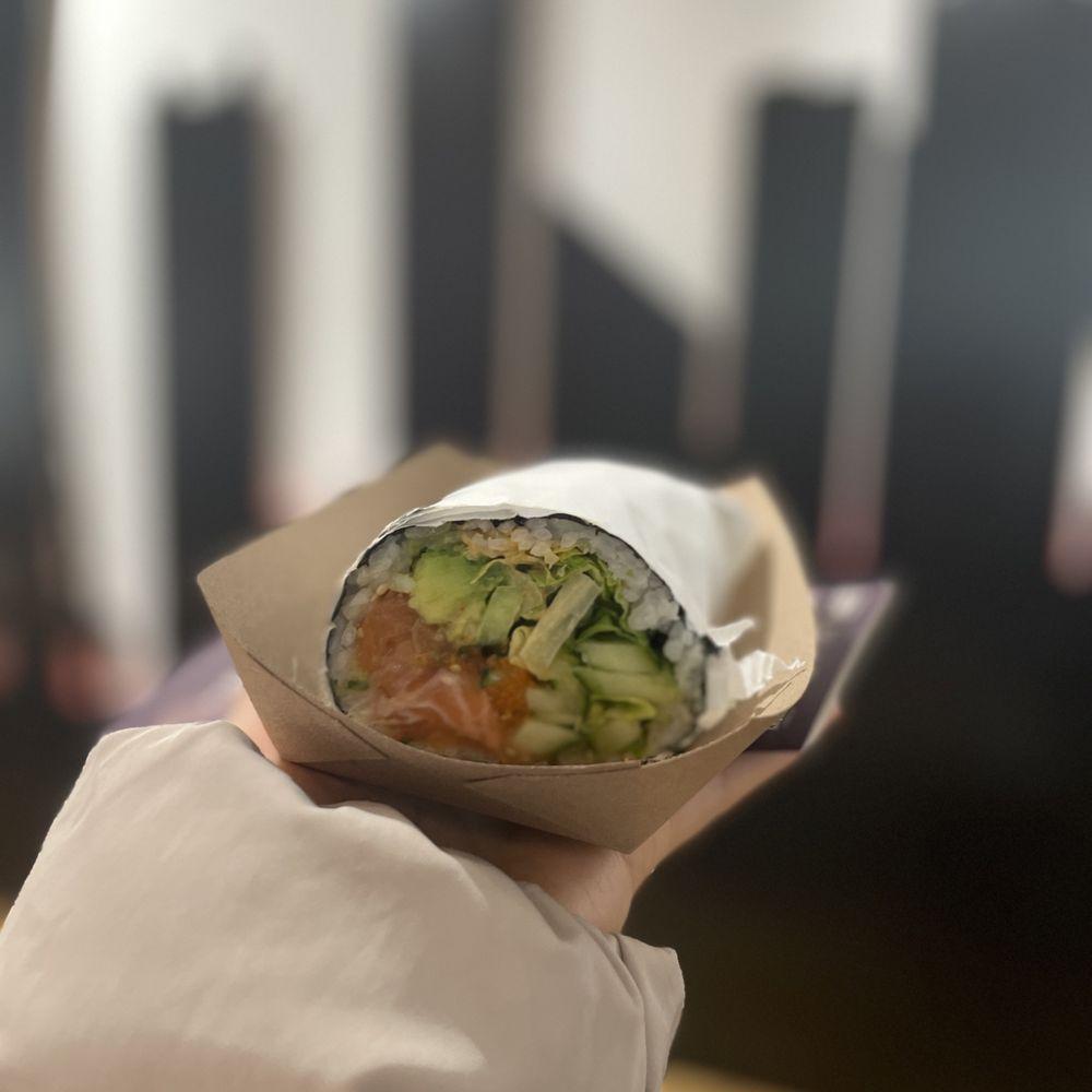 A person smiling while holding a Cucumber Cascade Sushi Burrito.