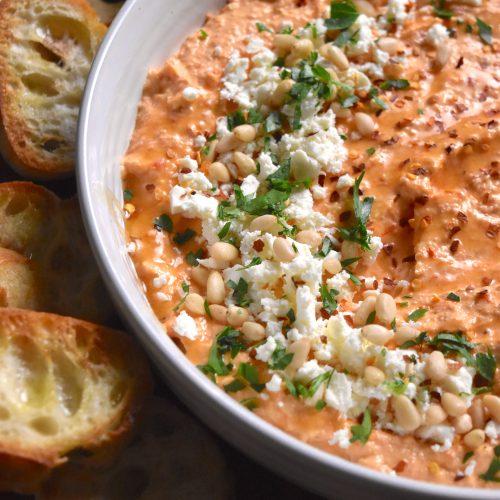 A vibrant bowl of roasted red pepper dip with feta cheese and olives, garnished with fresh herbs