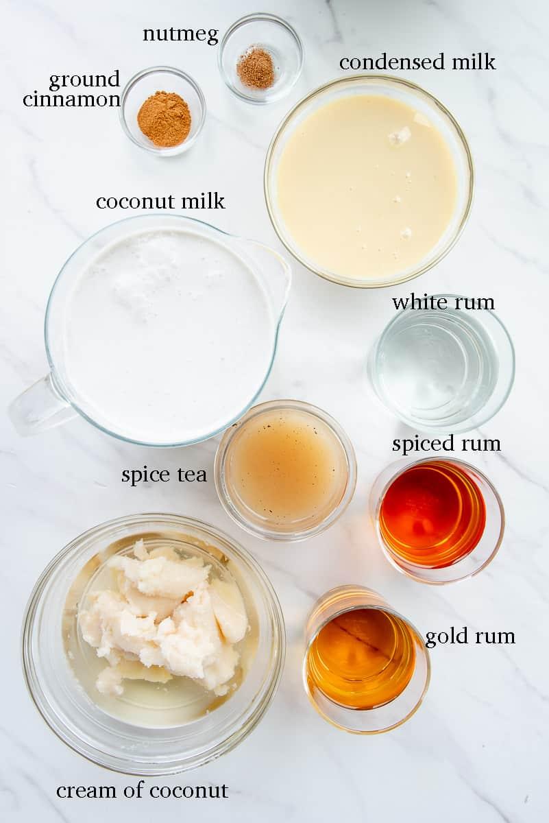 ingredients for coquito laid out: cans of coconut milk, cream of coconut, evaporated milk, condensed milk, rum bottle, vanilla, cinnamon sticks, nutmeg