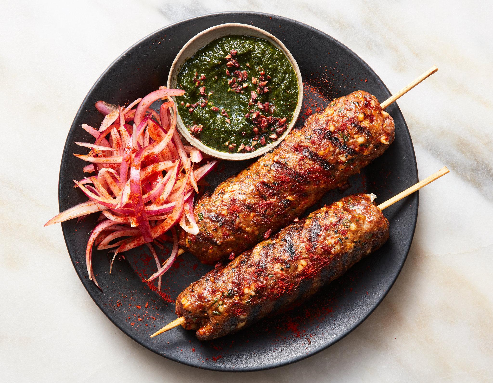 Close-up shot of sizzling lamb seekh kebabs on a grill, with a bowl of vibrant mint-cilantro chutney on the side