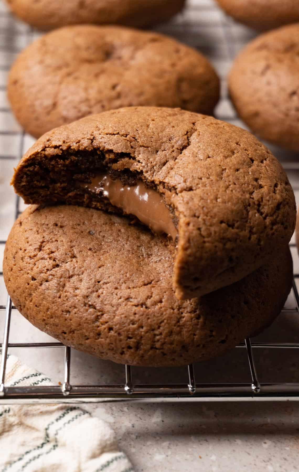 Warm, gooey Nutella-stuffed cookie with a bite taken out, showing melted Nutella center, on a cooling rack with more cookies in soft focus background