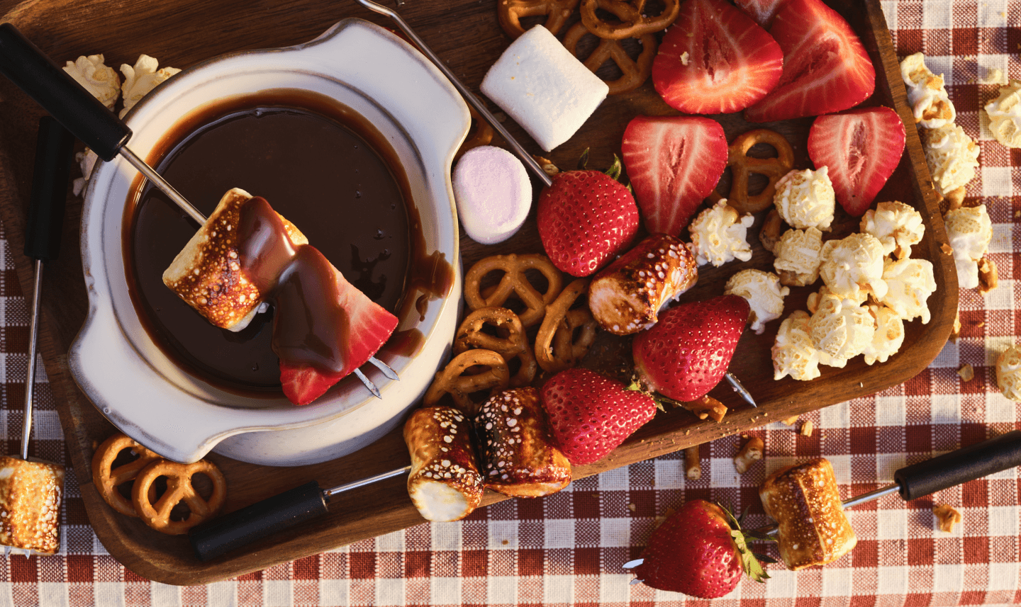 Close up shot of melted chocolate fondue with various dippers like strawberries, marshmallows, and cake pieces arranged around it.