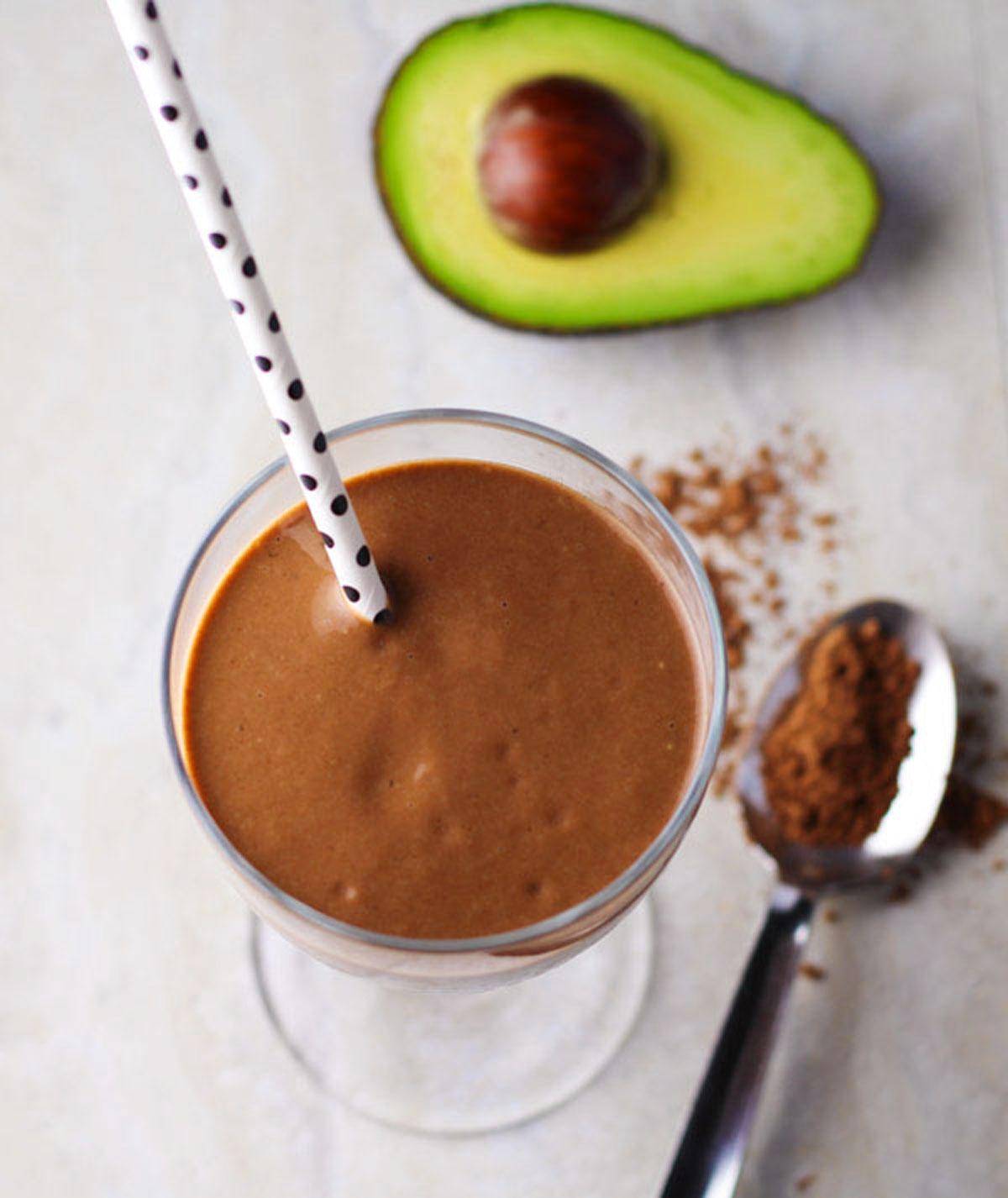 a chocolate avocado smoothie next to some fresh avocados and cocoa powder