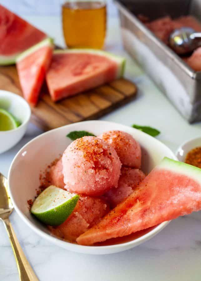 ingredients for watermelon sorbet laid out on a kitchen counter: watermelon chunks, lime, honey, and mint leaves