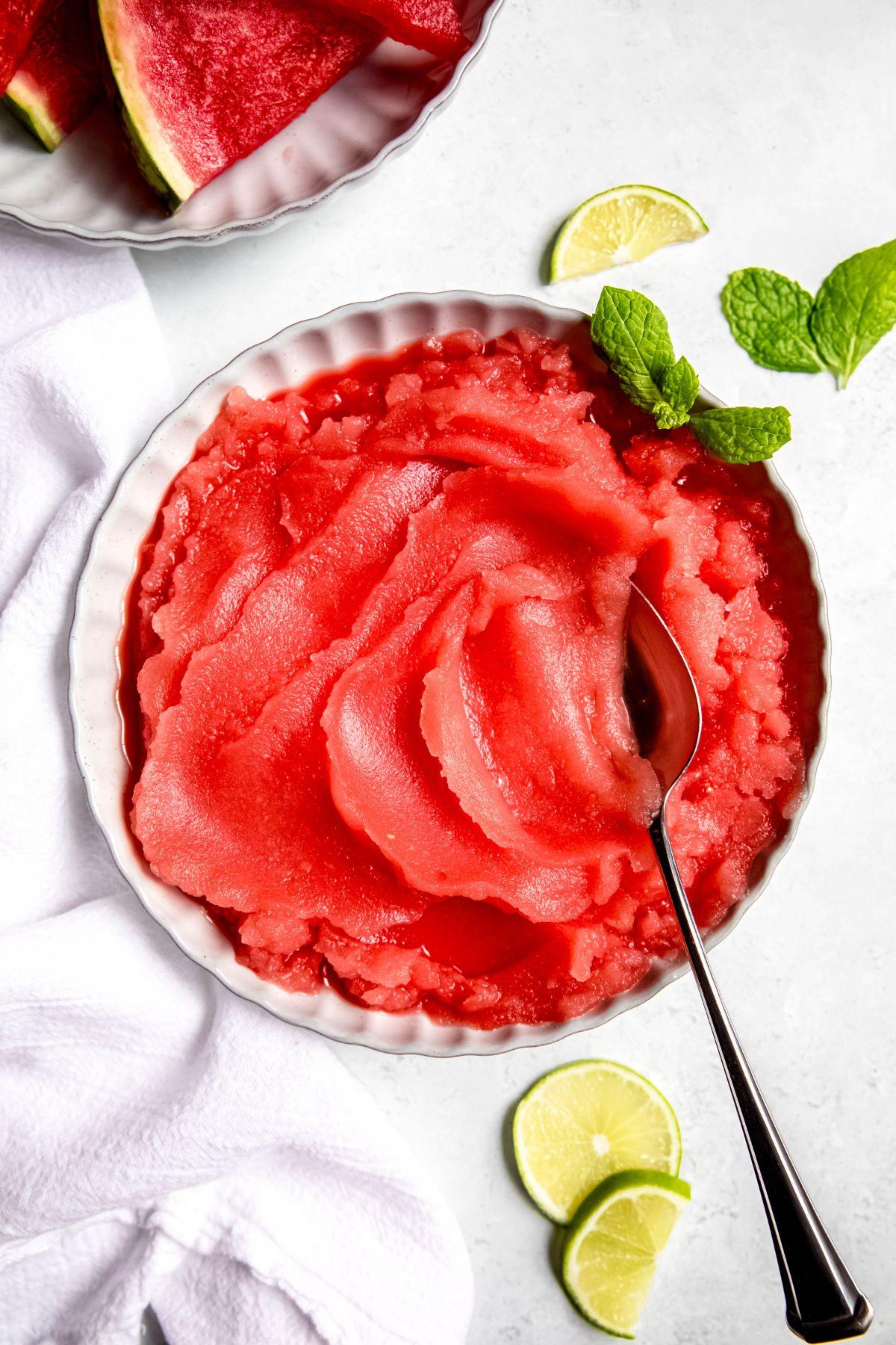 scoops of watermelon sorbet in a glass bowl, garnished with mint leaves and small watermelon wedges, against a bright, sunny background