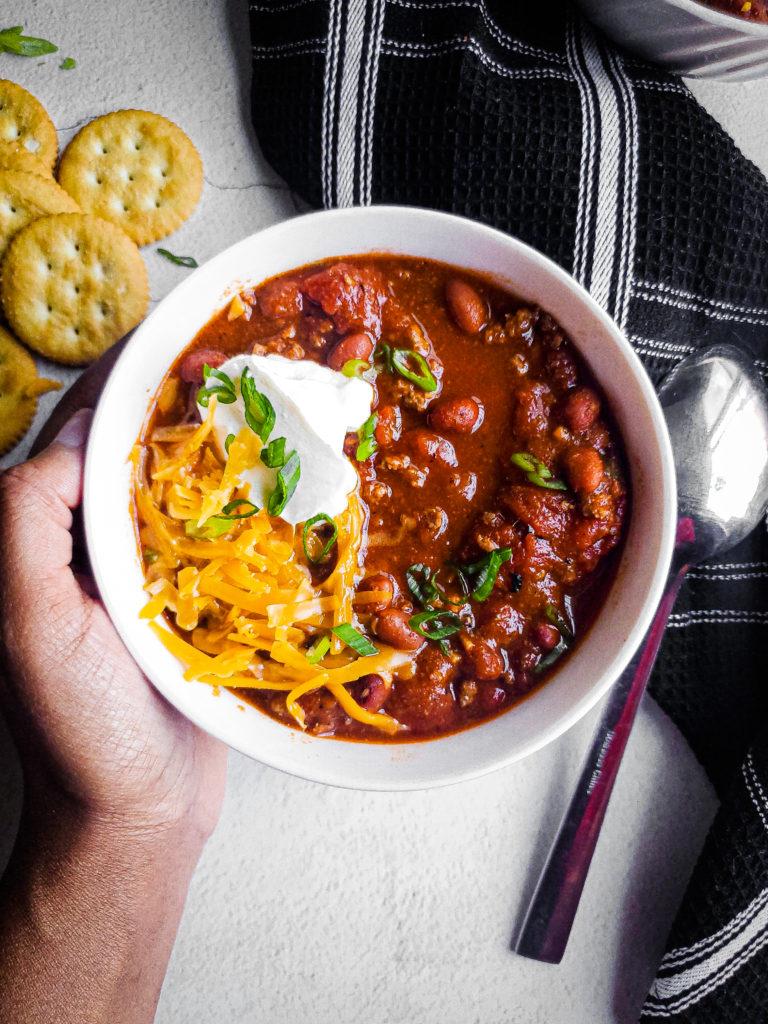 A hearty bowl of spicy beef chili topped with toasted pepitas, garnished with cilantro and a dollop of sour cream.