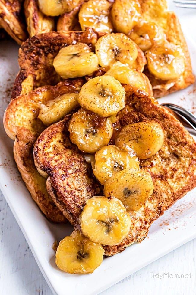 Close-up shot of Banana Foster French Toast drizzled with sauce and served with a side of whipped cream