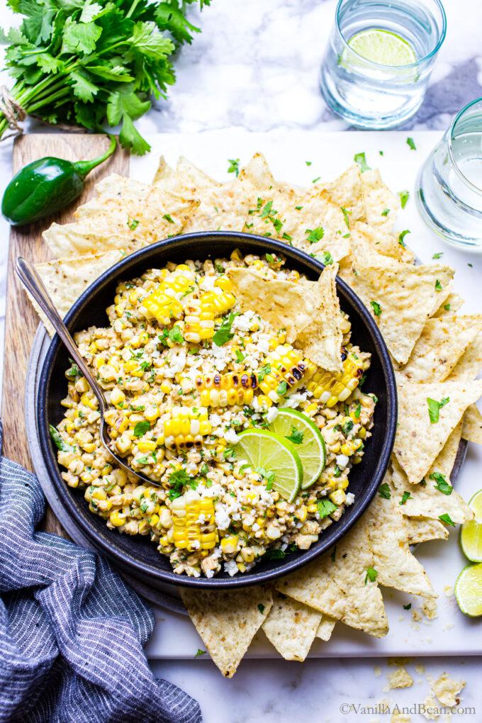 A bowl of Mexican street corn dip, garnished with cilantro and lime wedges, surrounded by tortilla chips