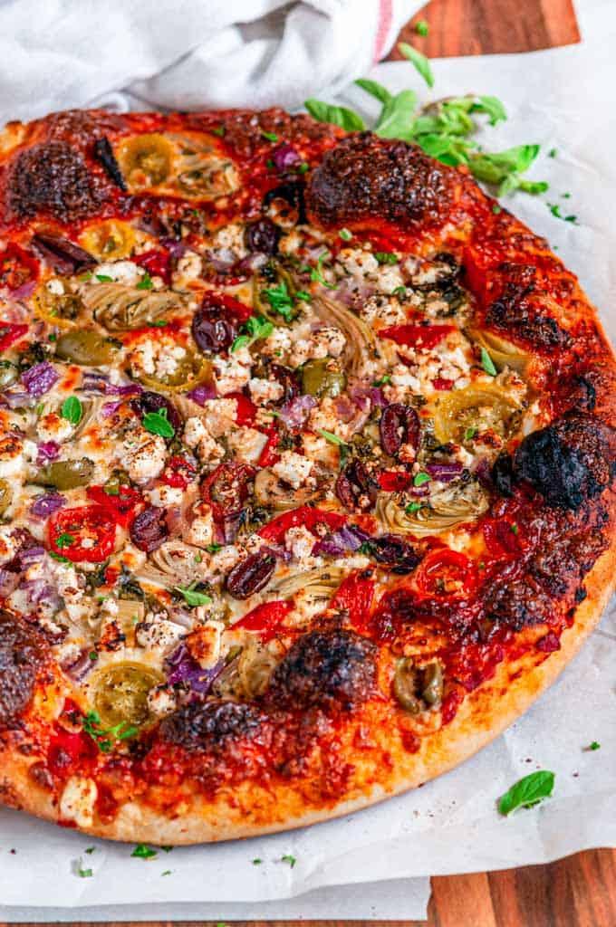 A whole pizza with olives and feta, served on a wooden board, garnished with fresh herbs