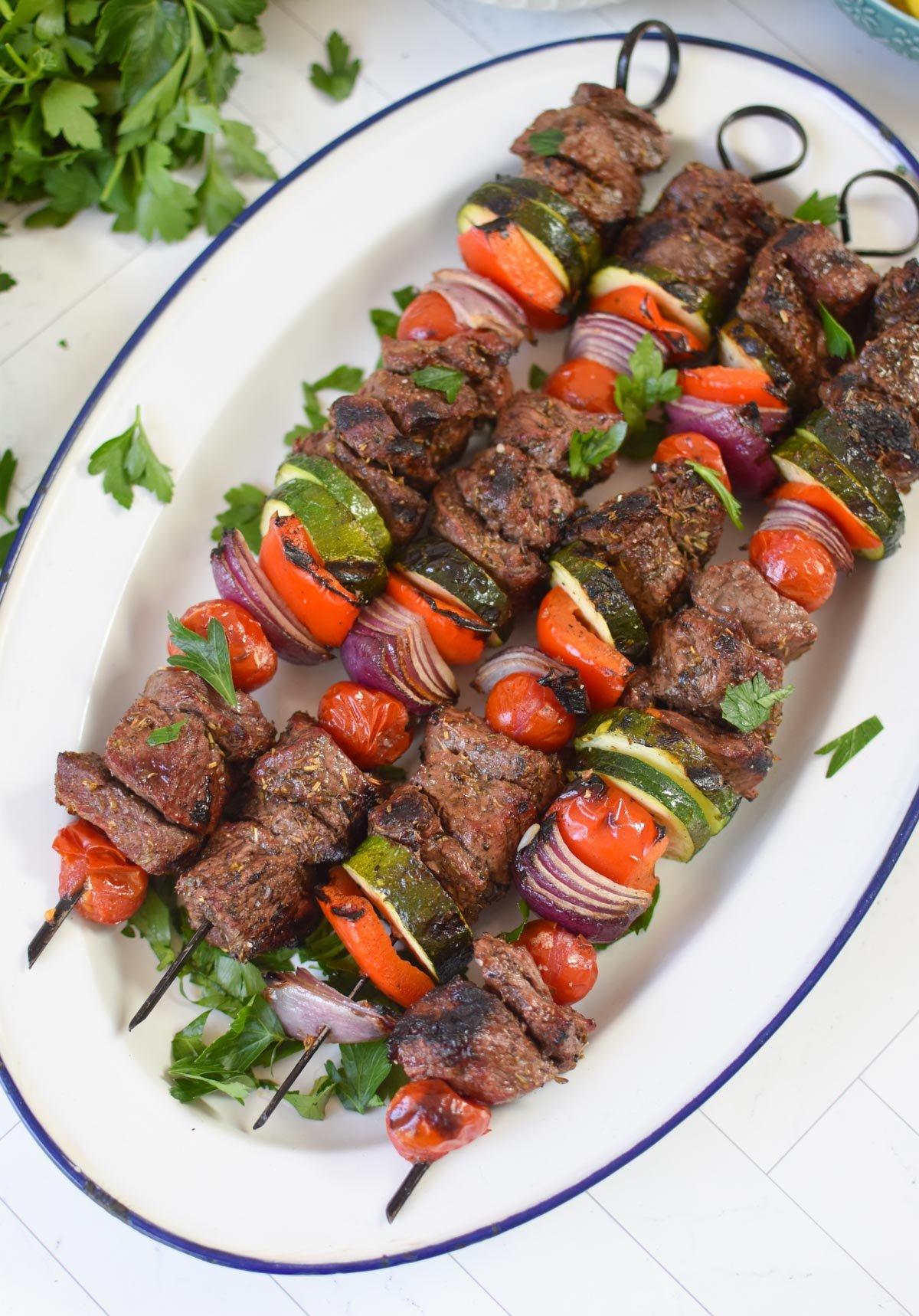 A platter of perfectly grilled beef souvlaki skewers arranged artfully with red onion marmalade on the side, garnished with fresh parsley.