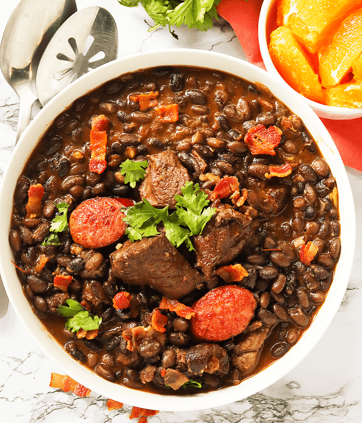 A steaming pot of Brazilian Feijoada with various meats and vegetables, garnished with orange slices and farofa.