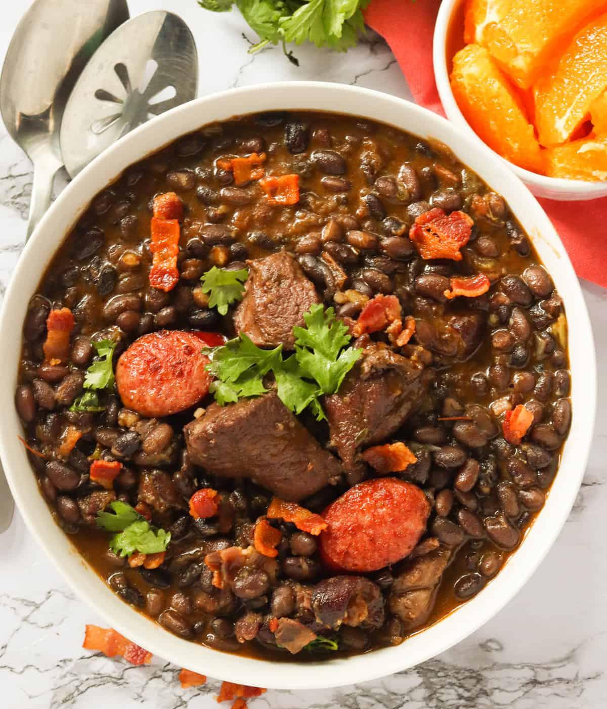 A hearty bowl of Feijoada with smoked ham hock and collard greens, garnished with orange slices and farofa.