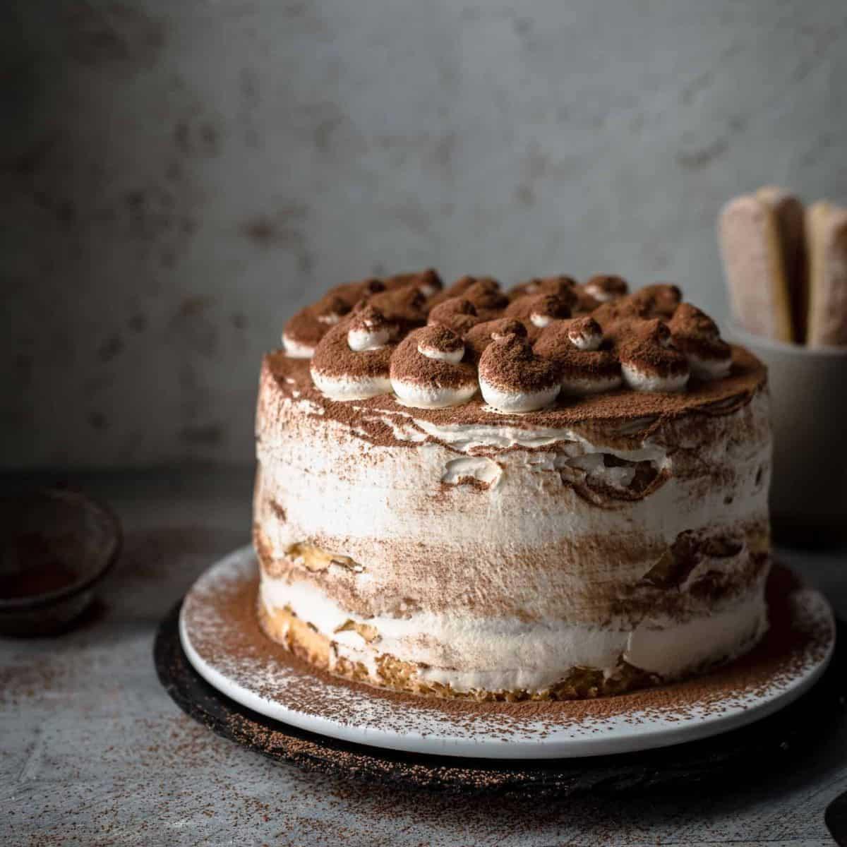 A whole tiramisu birthday cake sitting on a cake stand, garnished with cocoa powder and fresh berries