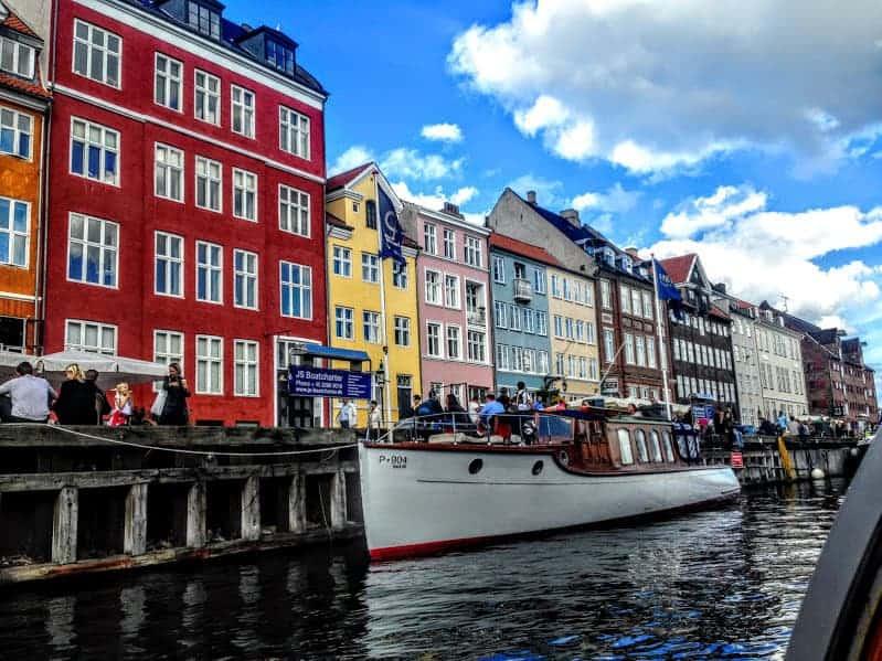 A young girl and her grandfather smiling, standing in front of a canal in Copenhagen, Denmark, with colorful buildings in the background.
