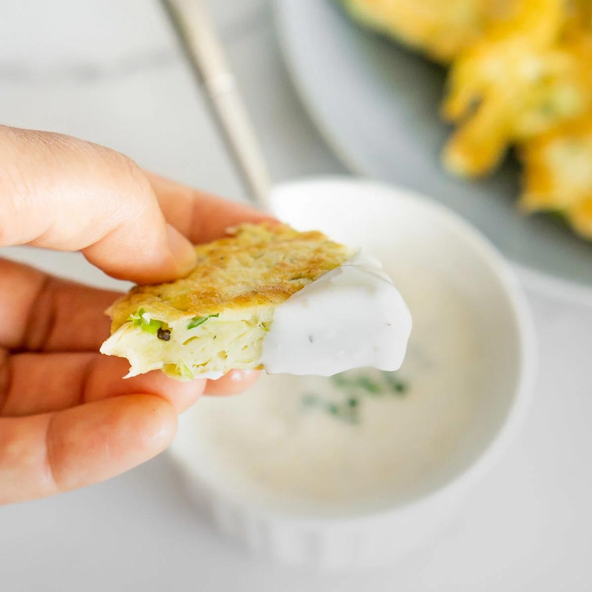 A hand dipping a fritter into a creamy dipping sauce