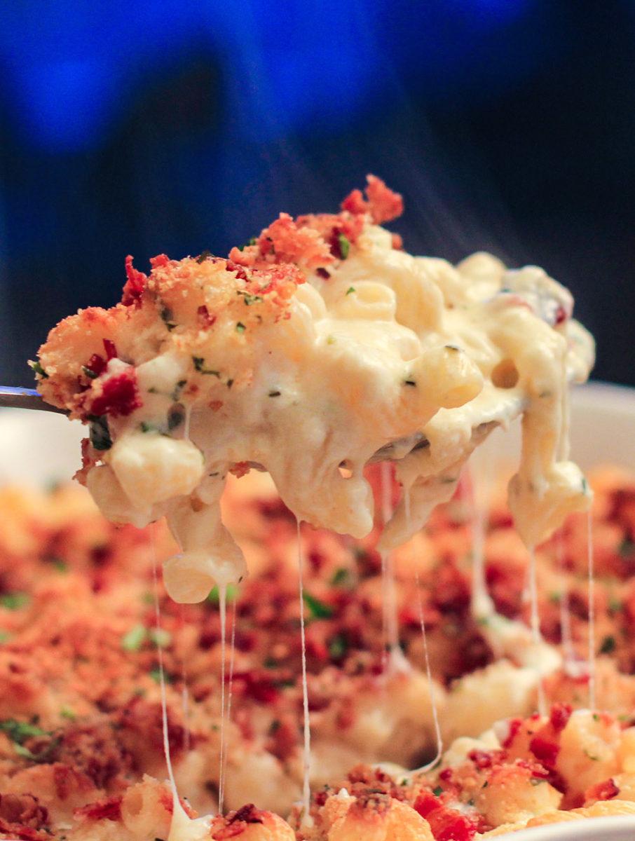 Close up shot of a creamy macaroni and cheese dish, topped with bacon bits and a swirl of ranch dressing.