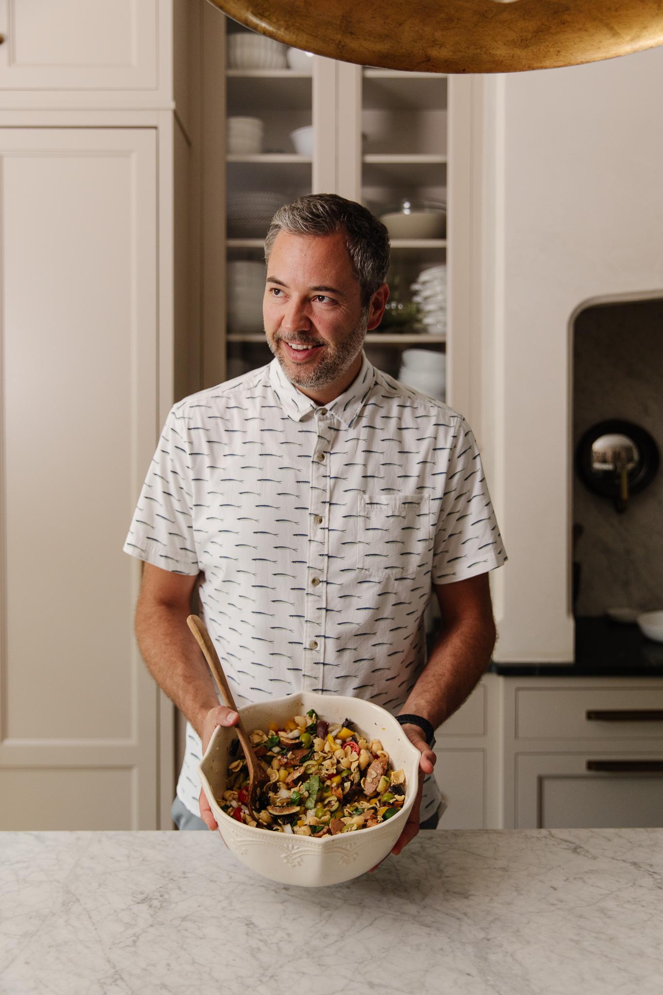 a person tossing the wheat pasta salad with a spoon in a brightly lit kitchen