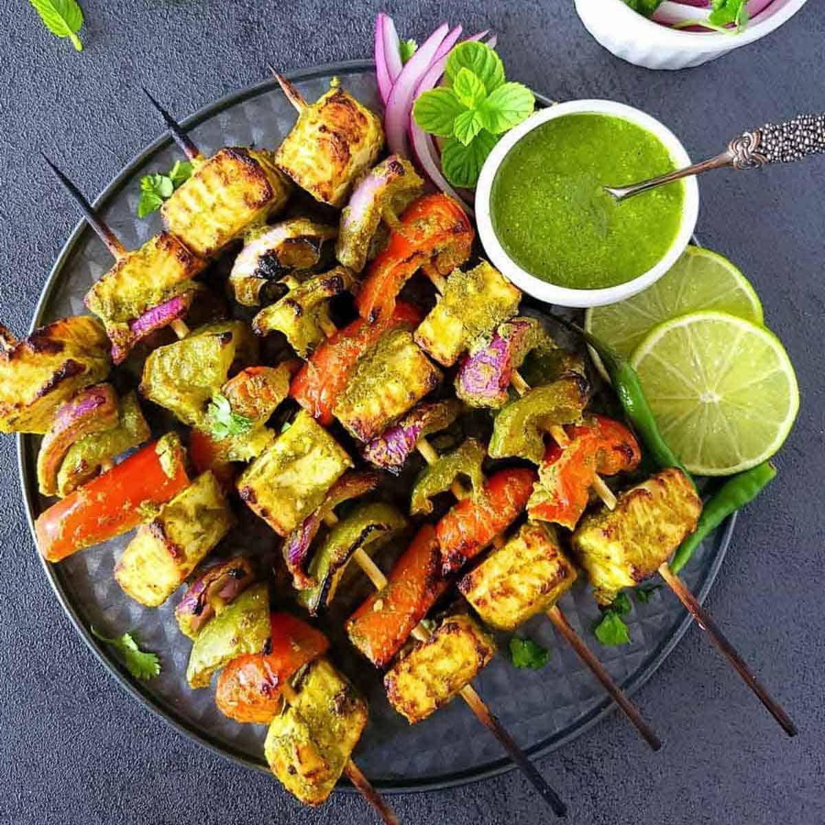 A platter of beautifully grilled tandoori paneer tikka, glistening with spices, served alongside a bowl of vibrant green mint chutney