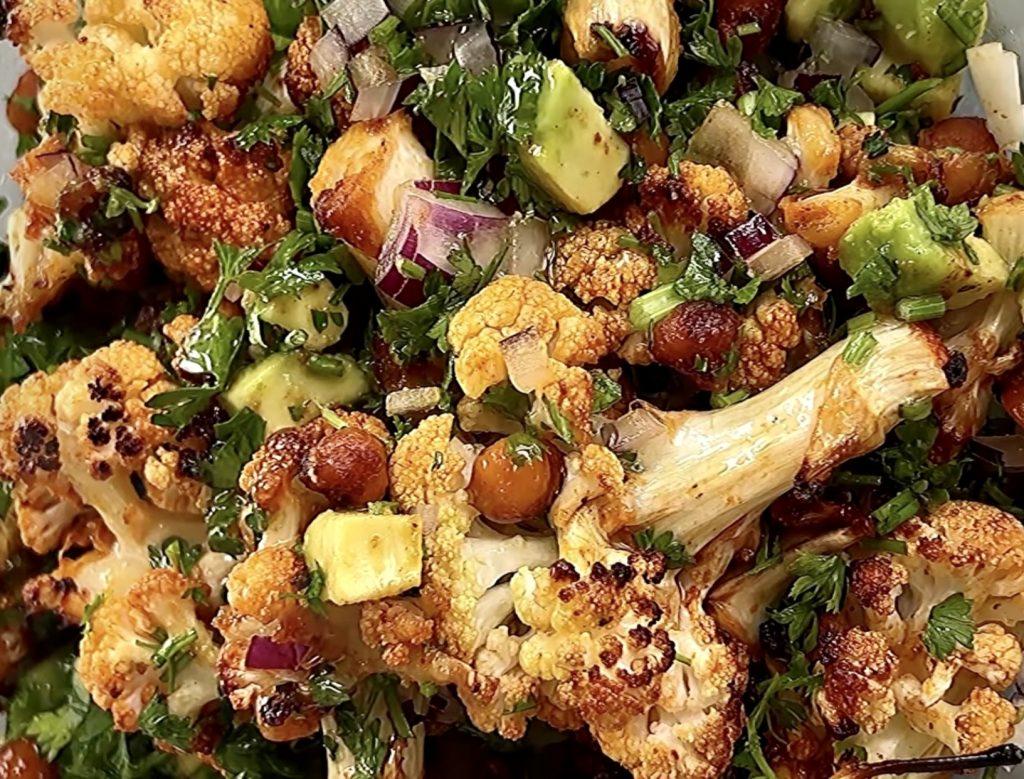 A rustic wooden table with a bowl of paprika-roasted cauliflower rice and other healthy ingredients, such as grilled chicken or chickpeas, arranged around it.
