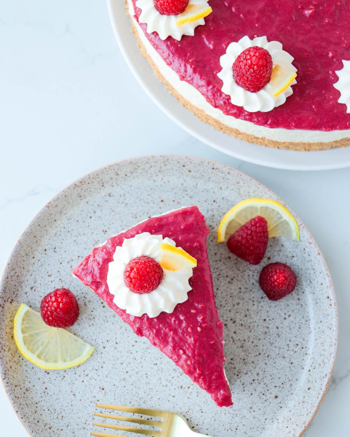 A slice of Lemon & Raspberry Cheesecake on a white plate garnished with fresh raspberries and lemon zest