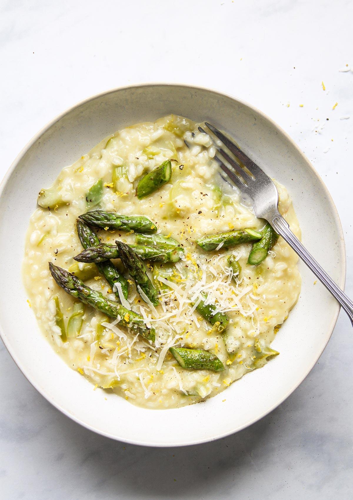 Overhead shot of a creamy asparagus risotto in a white bowl, garnished with parmesan crisps and fresh asparagus spears, set on a rustic wooden table