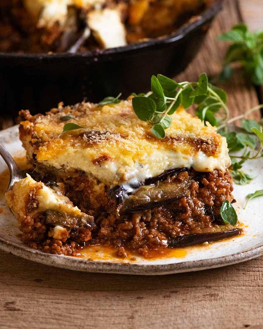 A wide shot of a Beef Moussaka baking dish, surrounded by fresh herbs and vegetables.