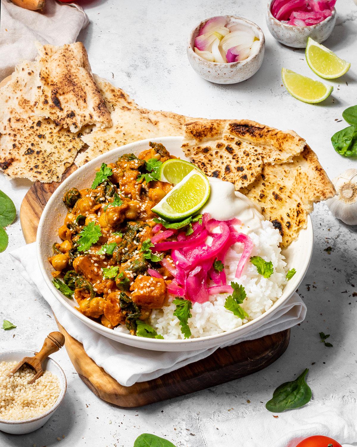 A rustic wooden table set with a bowl of spicy sweet potato and chickpea curry, rice, naan bread, and a glass of water