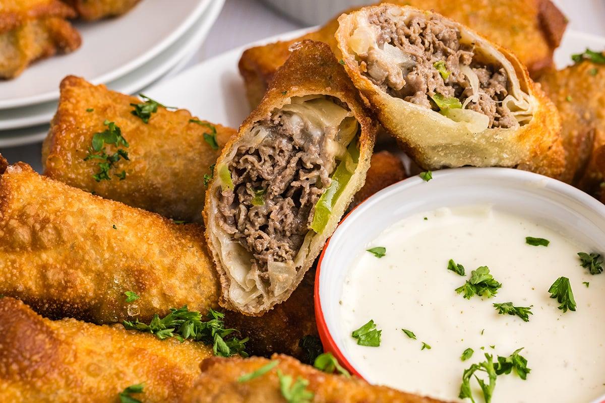 A platter of Philly Cheesesteak Egg Rolls, garnished with parsley and served with dipping sauces.