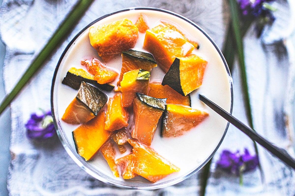 A person holding a glass of Kabocha Squash and Coconut Milk Smoothie, with a backdrop of autumn leaves and a cozy kitchen scene
