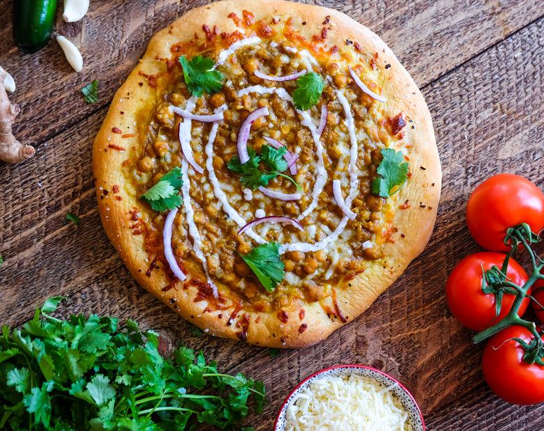 Overhead view of a whole Chana Masala Pizza with Green Chilies on a wooden cutting board, garnished with fresh cilantro